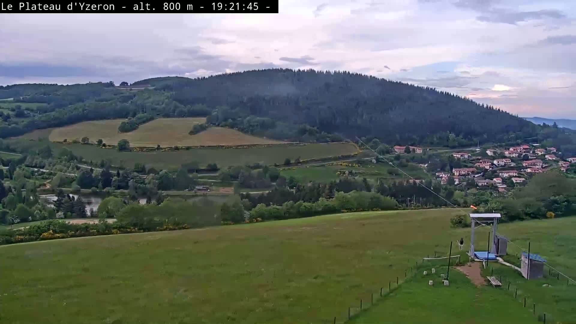 A high-angle, long shot shows a verdant landscape under a partly cloudy sky, featuring a small village nestled in a valley below forested hills, with a zip line in the foreground.