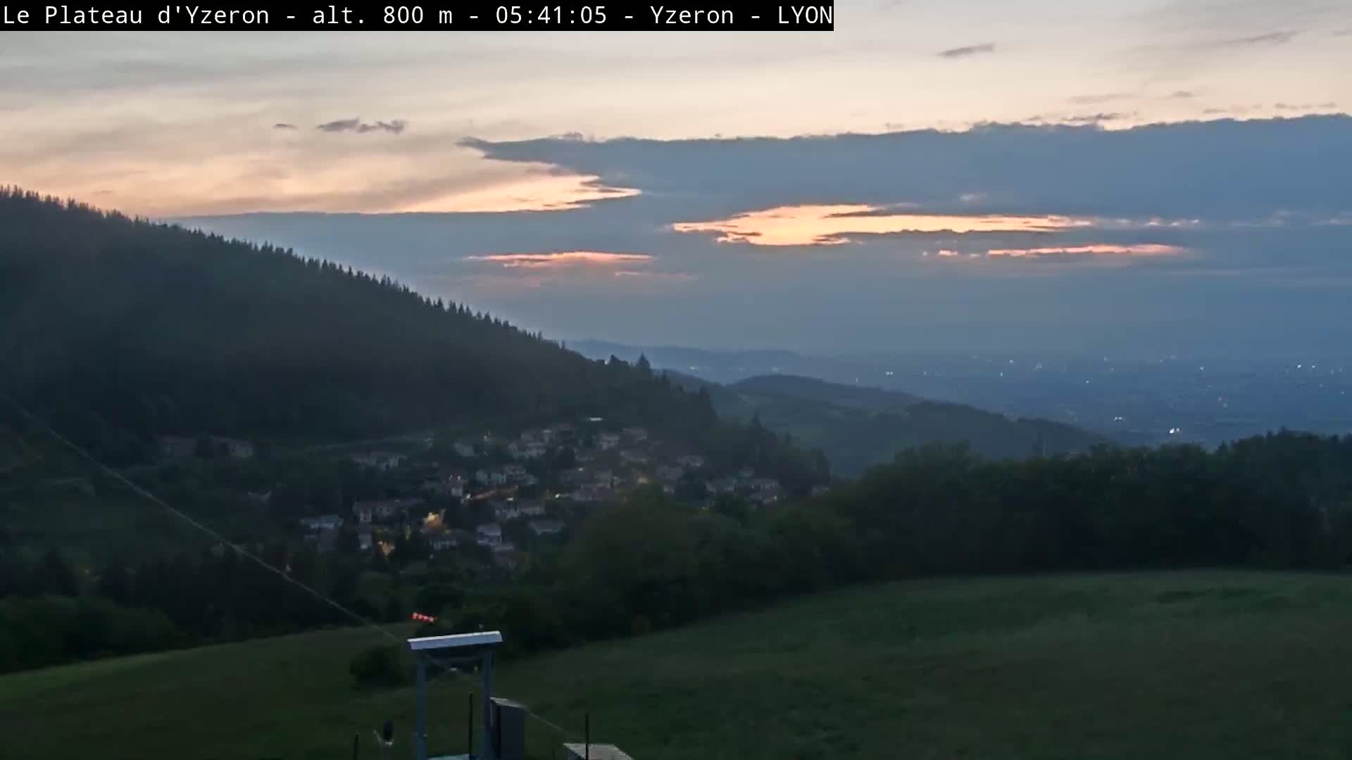 A twilight view shows a small town nestled in a valley below a hill covered in trees, under a partly cloudy sky with hints of orange in the clouds.