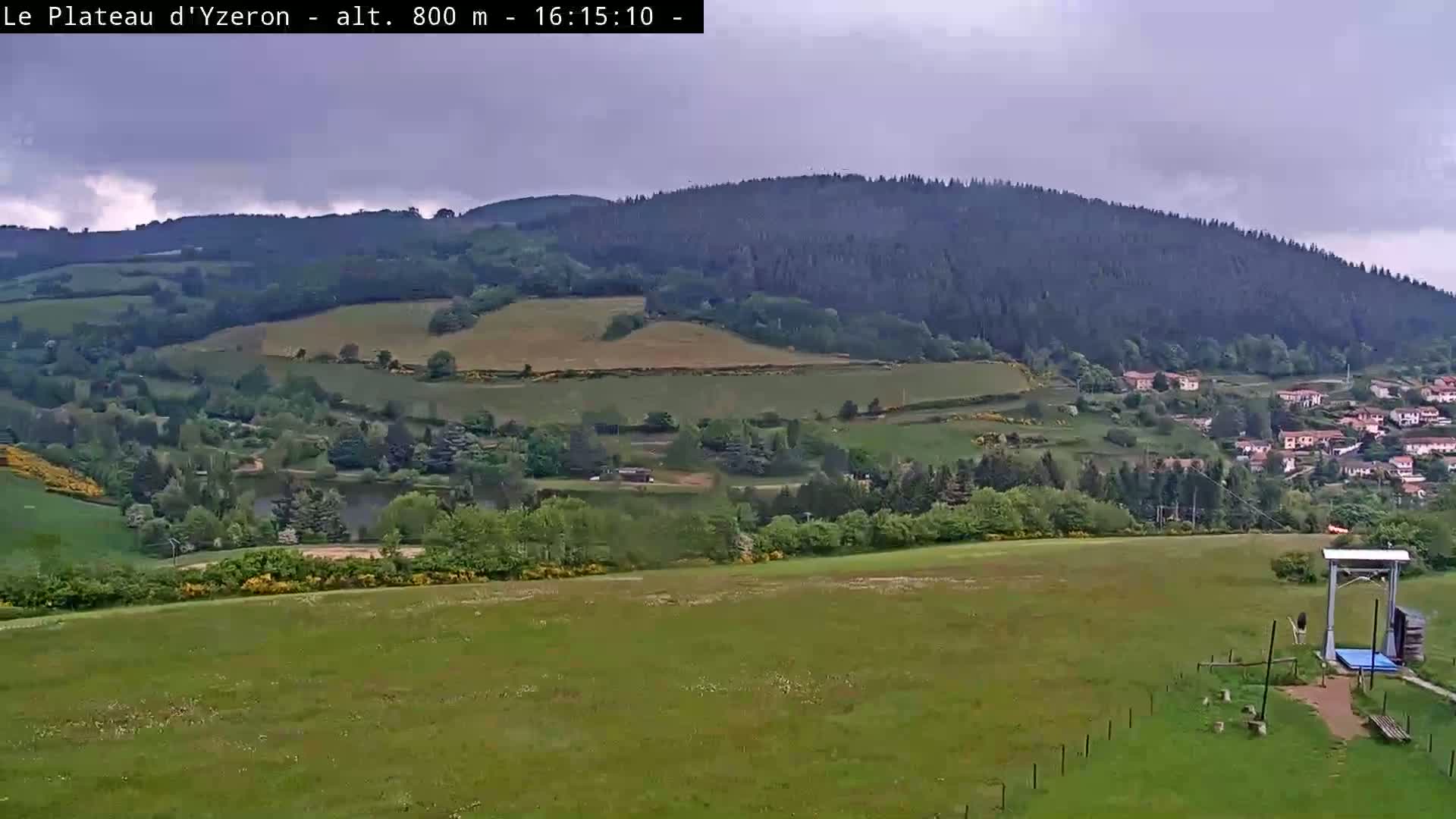 A grassy field overlooks a valley with a small lake, a village nestled among green hills, and a large forested mountain under an overcast sky.
