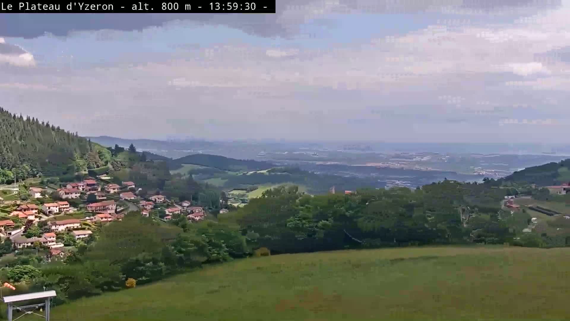 A partly cloudy day reveals a sprawling vista of a village nestled in a green valley, with rolling hills and distant plains stretching to a hazy horizon.