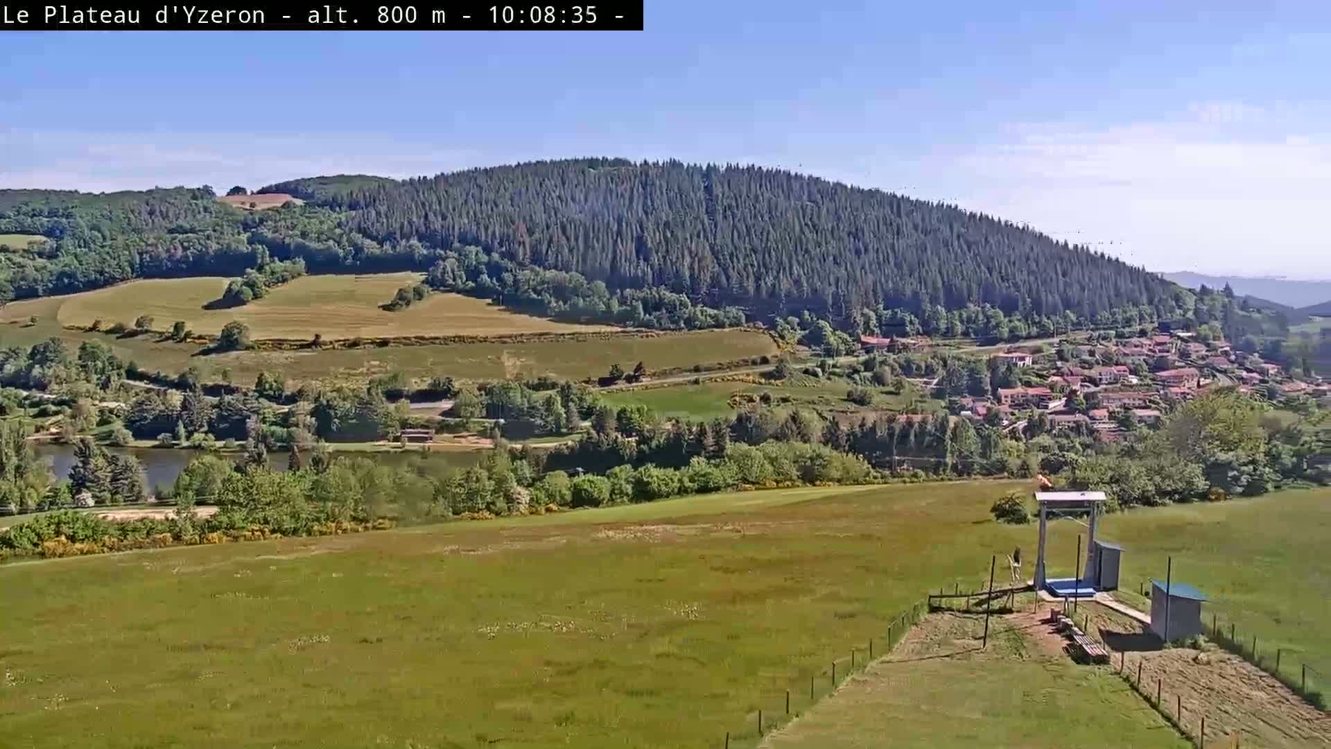 A sunny, high-altitude view of a verdant valley, featuring a small village nestled amongst fields and a forest-covered mountain in the background, with a ski lift visible in the foreground.