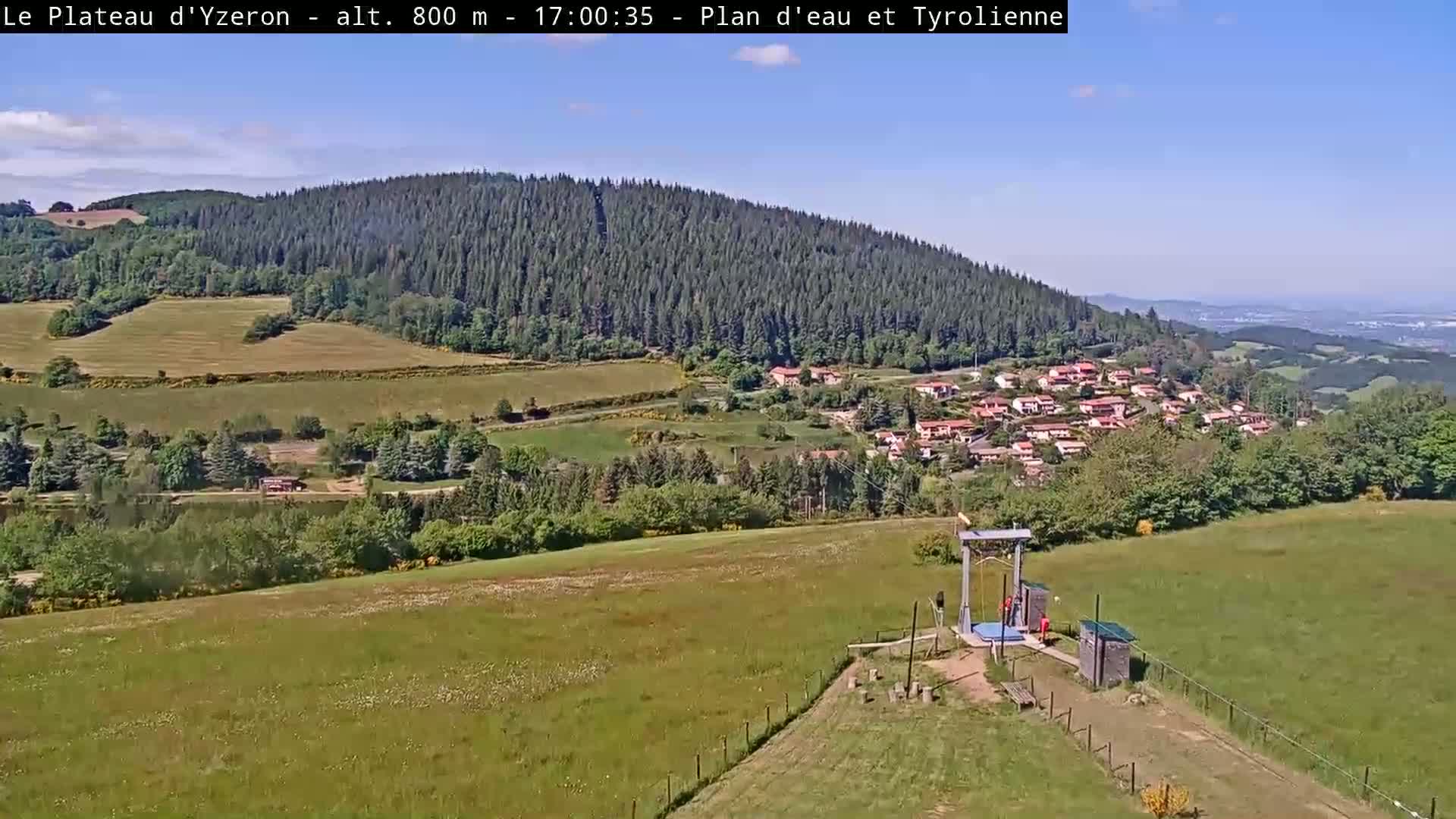 A sunny, outdoor view shows a grassy field with a small structure and a zipline, overlooking a village nestled in a valley below a forested mountain.