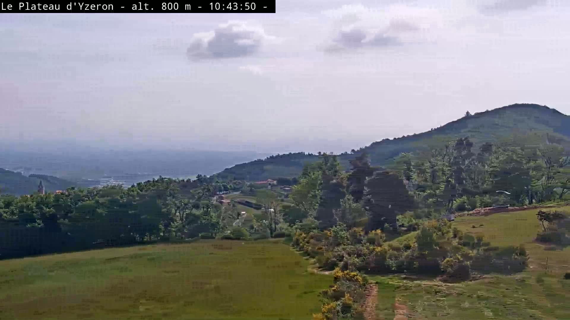 A partly cloudy day reveals a verdant landscape of rolling hills, lush green trees, and a distant valley under a hazy sky.