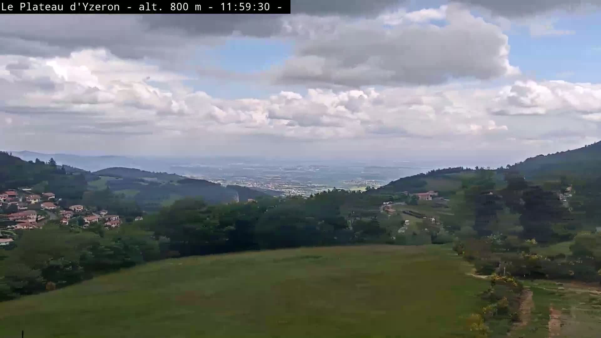A partly cloudy sky overlooks a sprawling green landscape dotted with houses and trees, extending to a distant valley.