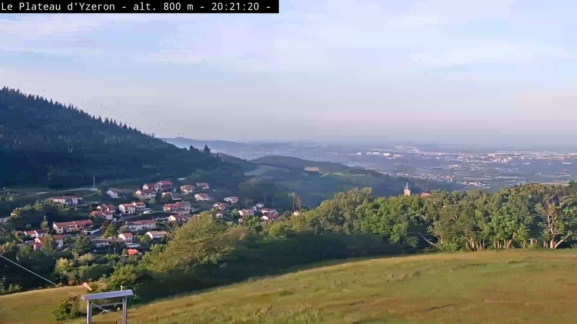 A hazy, long-range view shows a small village nestled in a valley, overlooked by forested hills and a distant cityscape under a clear sky.