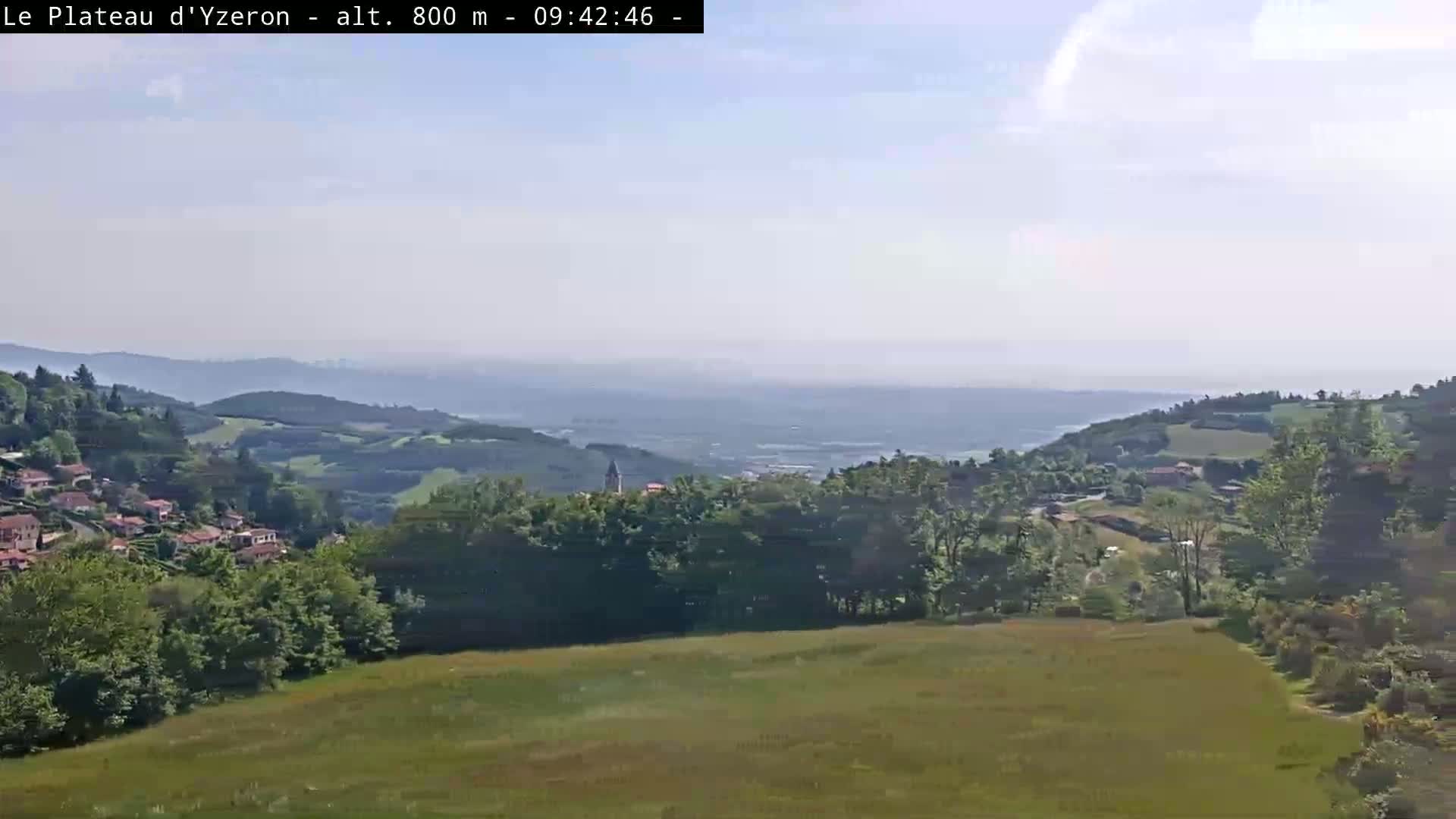 A hazy, sunny view of a verdant valley with a small village nestled among green hills, extending to a distant horizon.