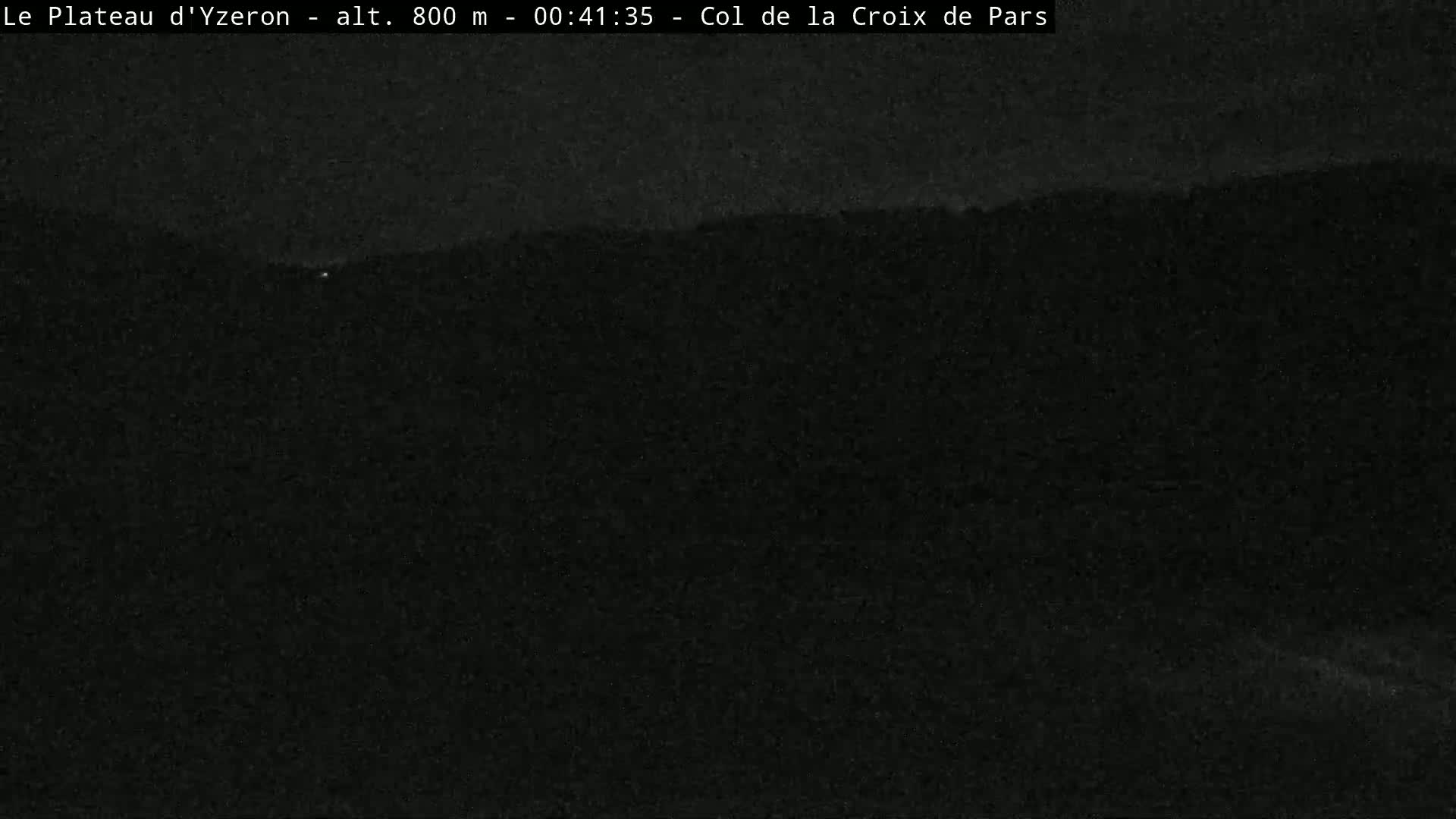 A nighttime, low-light image shows a dark, indistinct landscape of trees and hills under a dark sky.