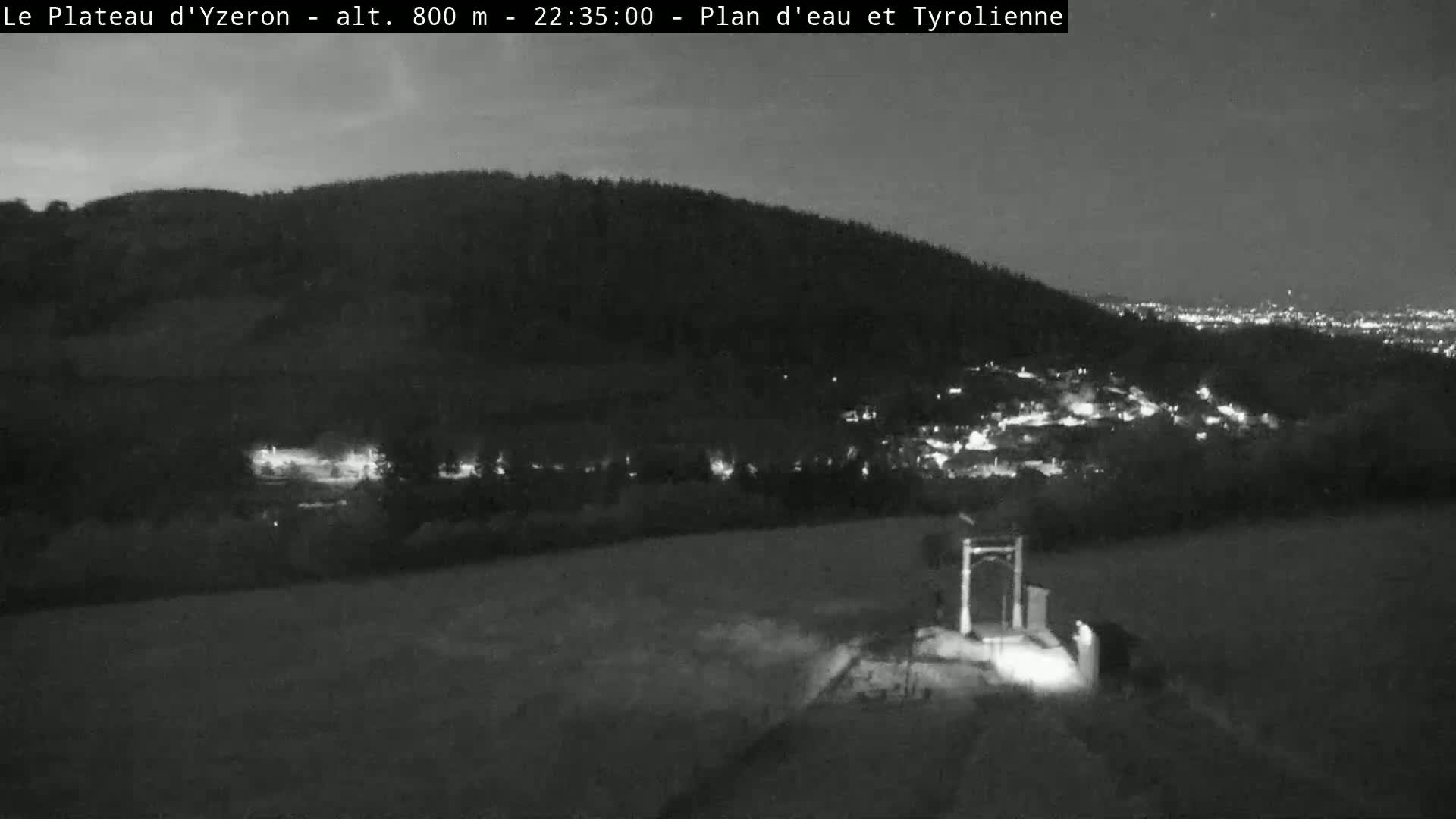 A nighttime, low-light view shows a dark hill overlooking a small town with scattered lights, under a mostly clear night sky.