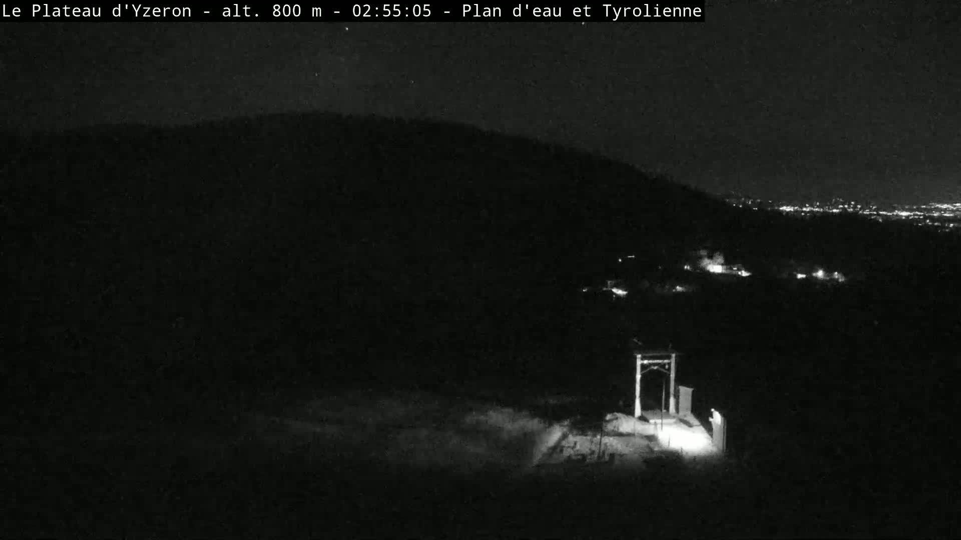 A nighttime, low-light view shows a dark hillside, a structure with lights near the bottom, and a distant city glowing faintly in the background.