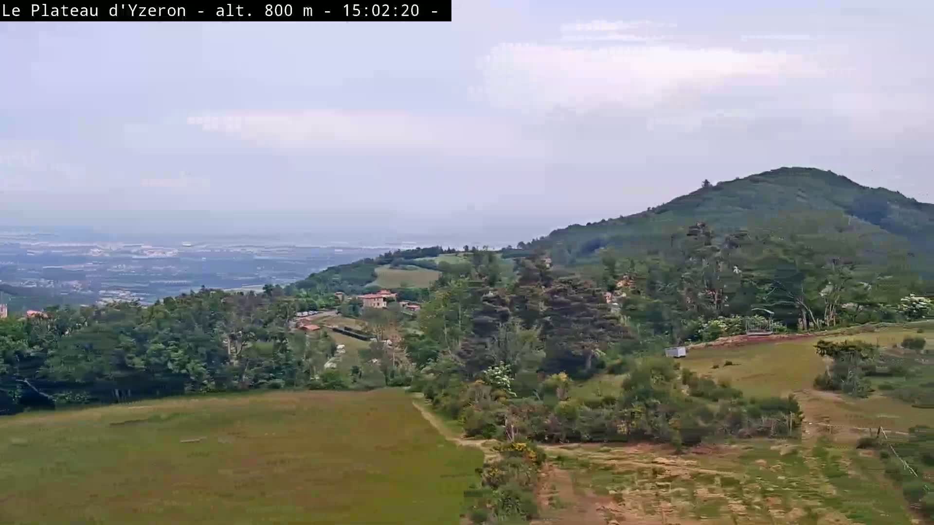 A mostly clear day reveals a verdant landscape of rolling hills, trees, and fields, overlooking a distant city and a hazy body of water.