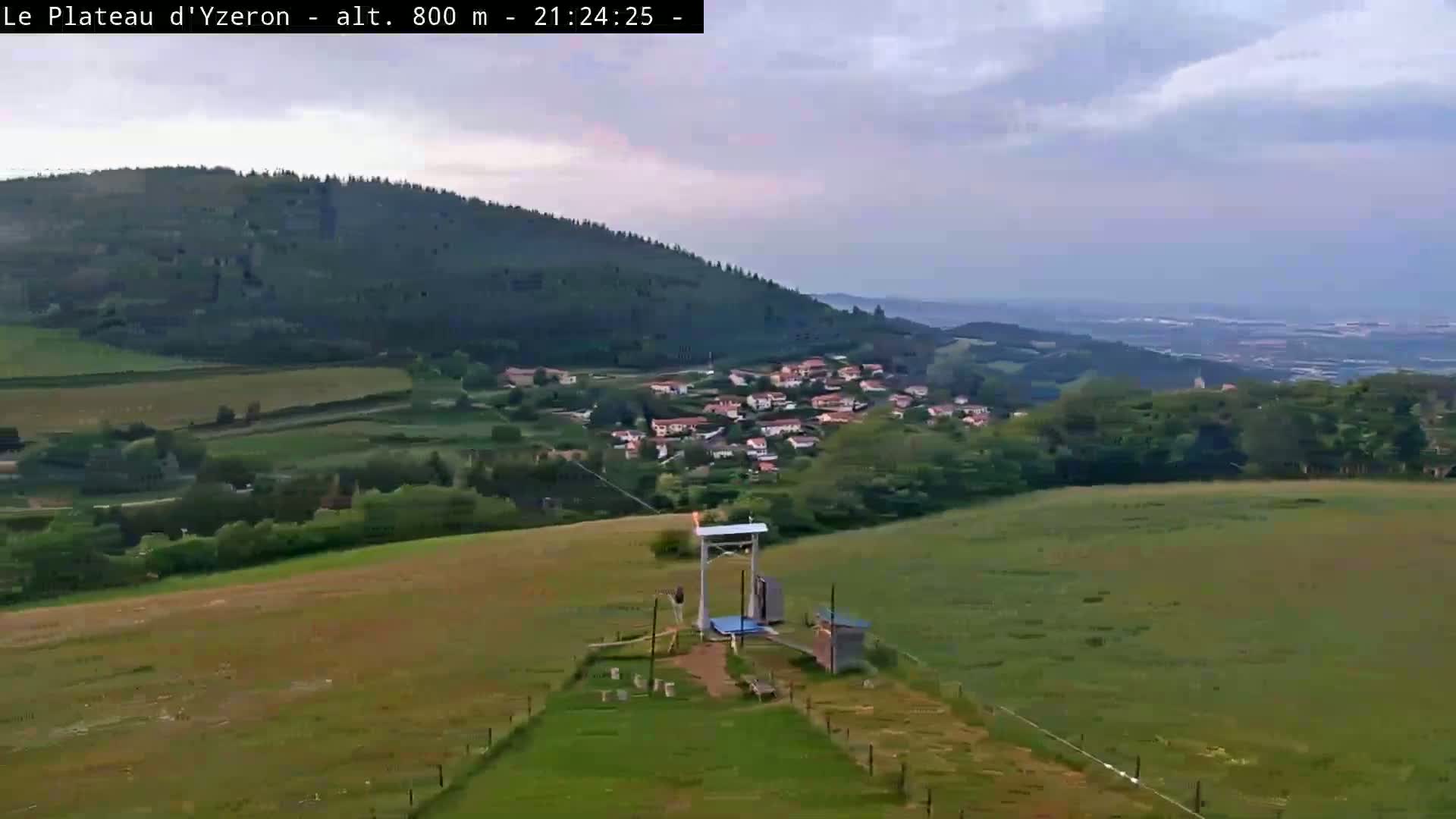 A mostly cloudy evening shows a small village nestled in a valley between rolling green hills, with a lift or ski-type structure in the foreground.