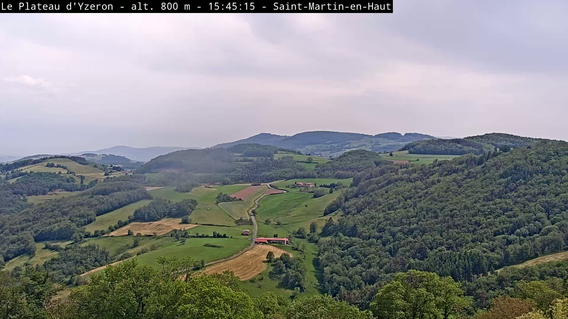 A partly cloudy, high-altitude view of a verdant valley dotted with farmhouses and fields, surrounded by rolling hills covered in lush green forests.