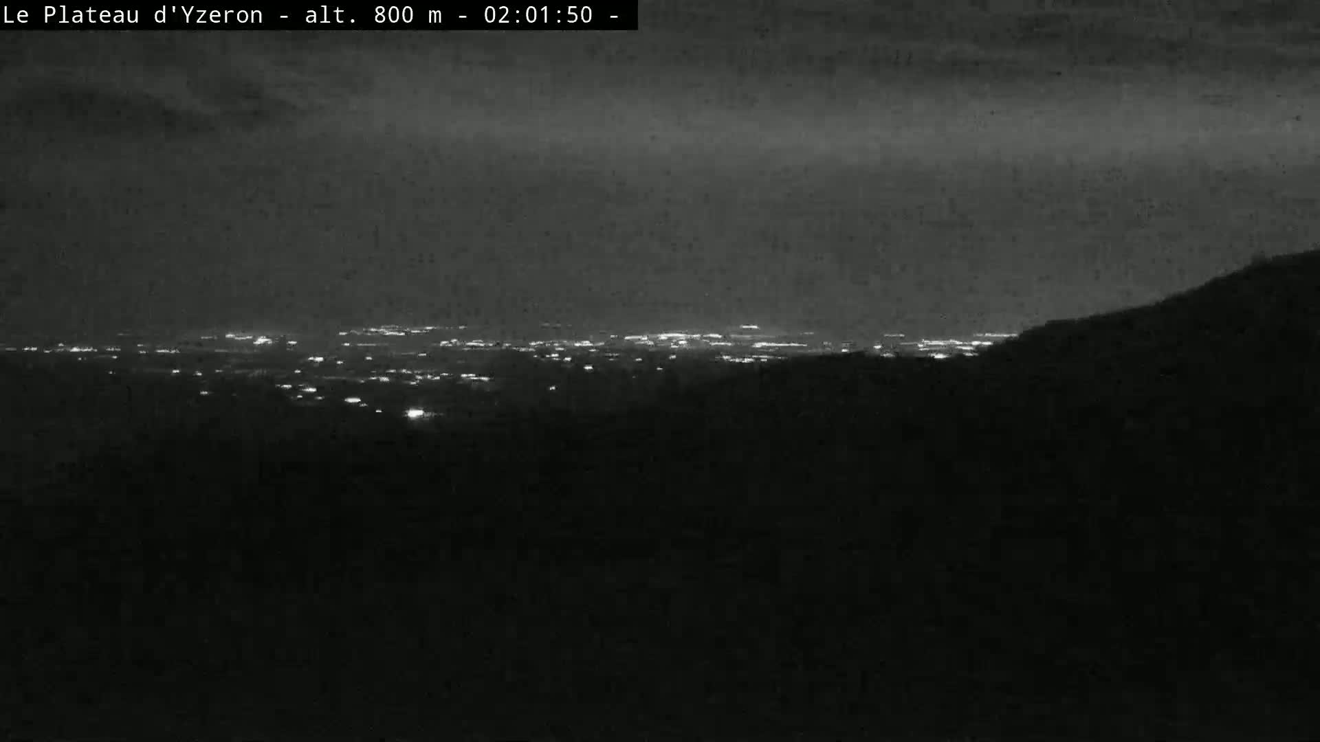A nighttime view from a hilltop shows a distant city glowing with lights under a dark, cloudy sky.