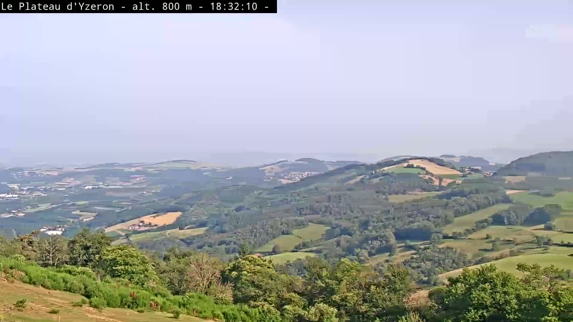 A hazy, long-range view of a verdant valley dotted with fields and forests under a light, clear sky.