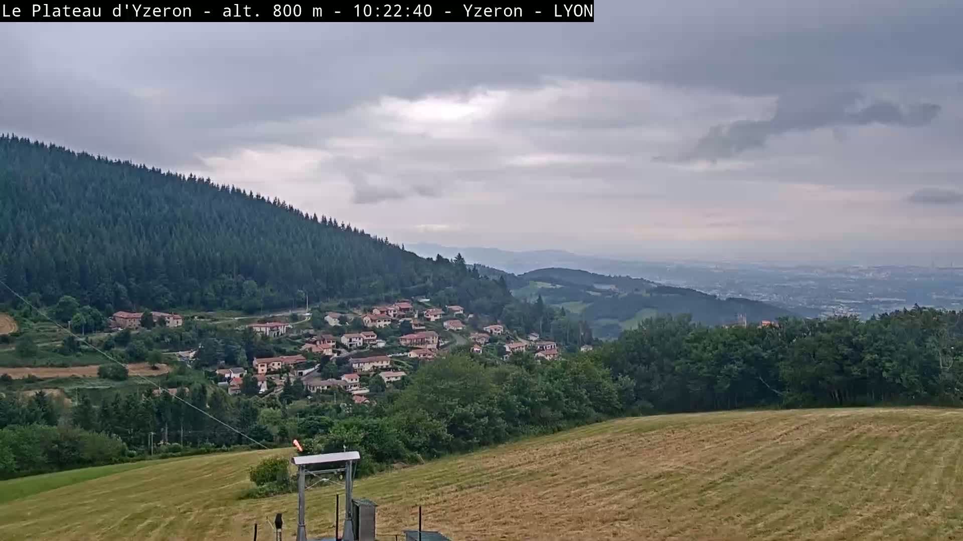 A hillside village nestled in a valley below a forested mountain, with a distant view of a city under a cloudy sky.