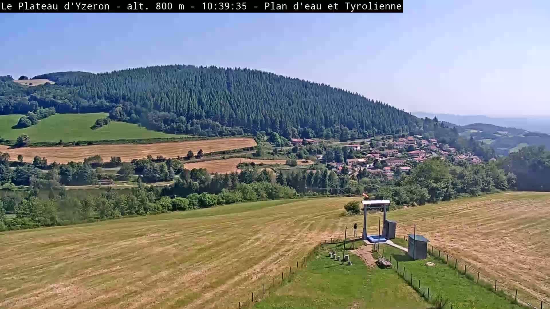 A sunny, high-altitude view shows a small village nestled in a valley surrounded by green fields and a large, dark green forest-covered mountain.
