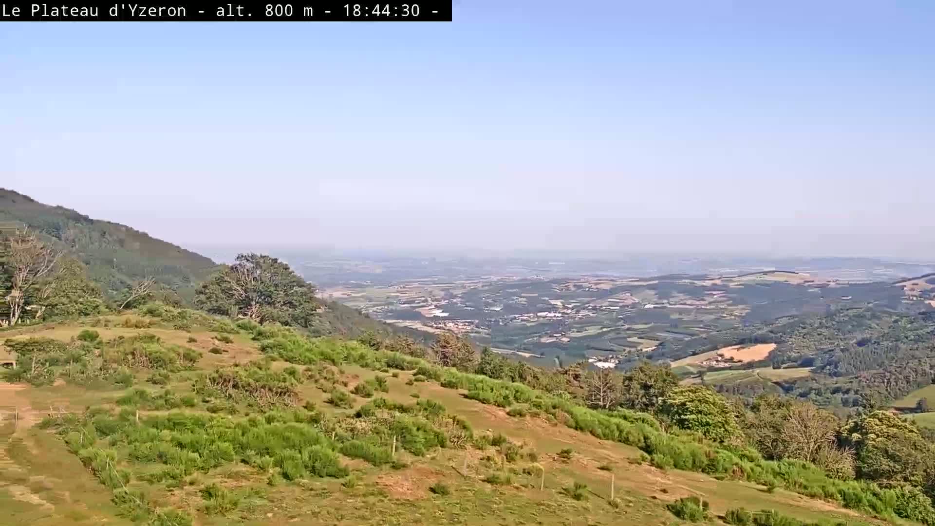 A vast, hazy valley spreads out below a grassy hillside on a clear day.