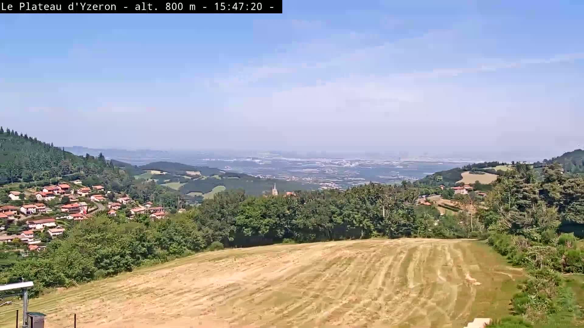 A hazy, sunny day reveals a vast panorama of rolling hills, a village nestled among green trees, and a distant, expansive plain stretching to a hazy horizon.