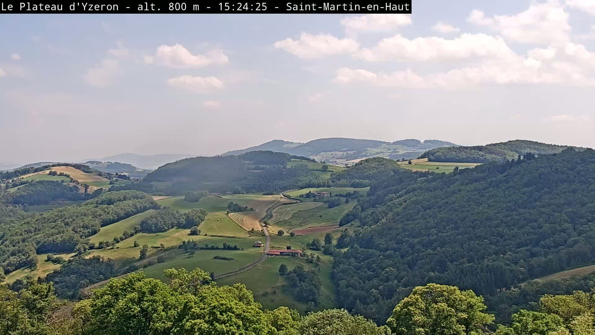 A partly cloudy sky overlooks a vast, rolling landscape of green hills, fields, and forests.