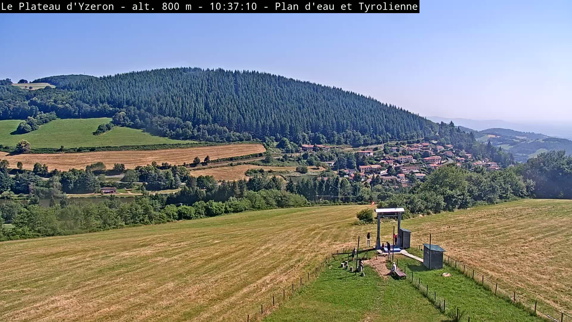 A sunny view shows a grassy field leading to a small village nestled below a large, forested hill, with a few people gathered near a structure in the foreground.