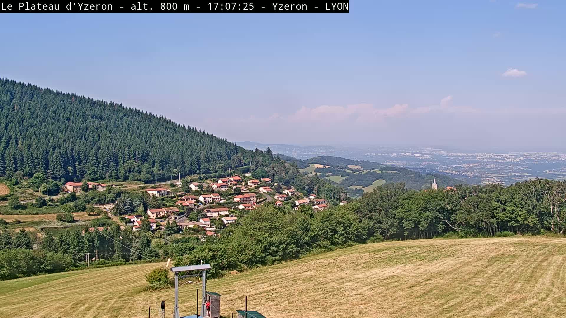 A hilly landscape under a mostly clear blue sky shows a village nestled among green trees and fields, with a distant view of a large city or town in the background.