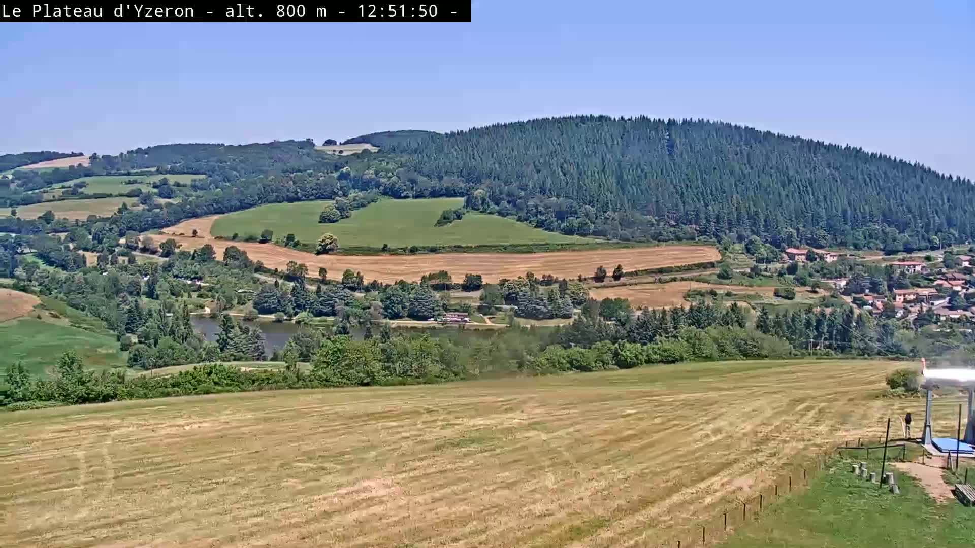 A high-angle view on a sunny day shows a vast expanse of farmland and forest, including a small lake and a village nestled in the valley.