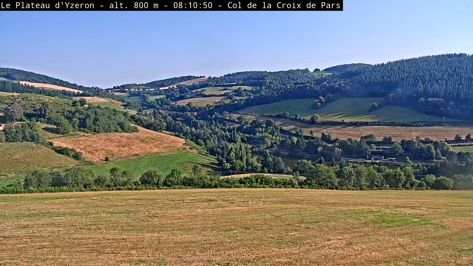 A sunlit valley dotted with green trees and fields of varying shades of brown and green stretches under a clear blue sky.