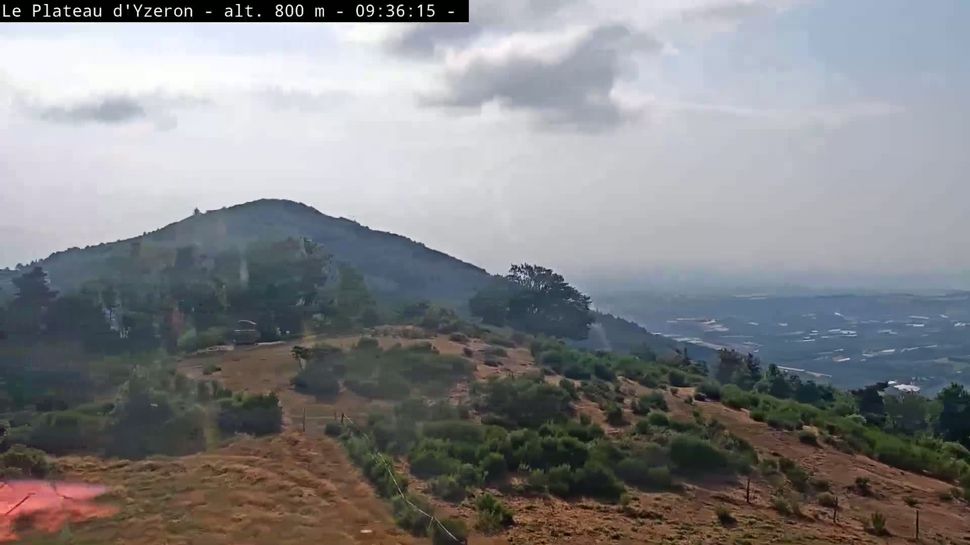 A hazy, partly cloudy day reveals a view of a dry, scrub-covered hillside and distant valley from a high vantage point.