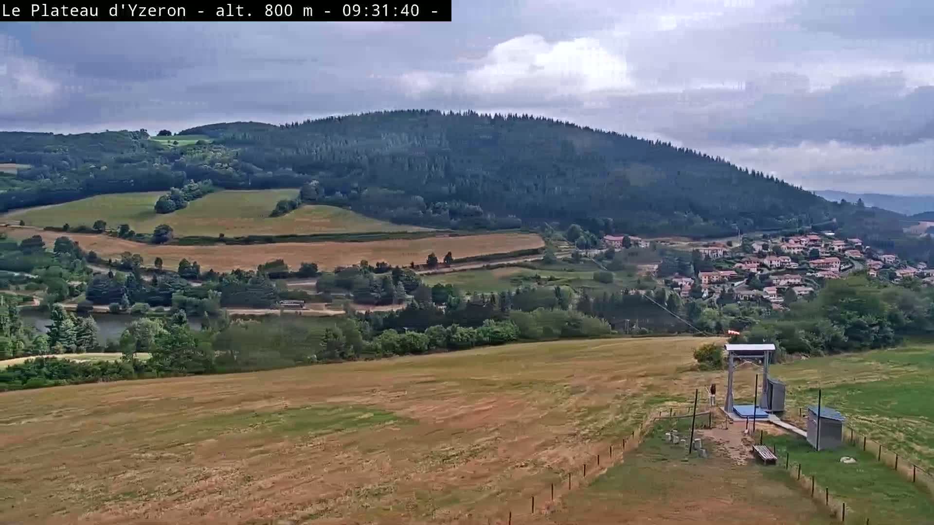 A high-angle view shows a partly cloudy day overlooking a valley with a small village, fields, and a small structure possibly related to a zipline or cable car.