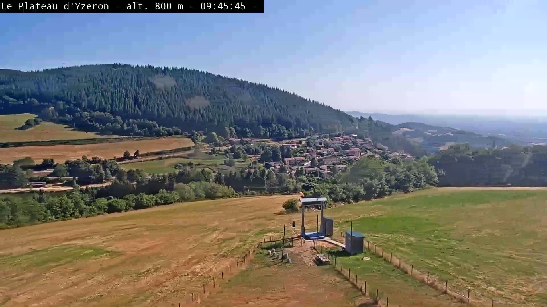 A sunny view from a hilltop shows a small village nestled in a valley surrounded by fields and forests, with a small structure and fenced area in the foreground.