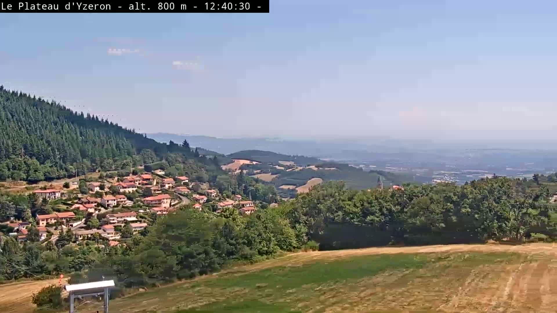 A sunny view from a hilltop shows a small village nestled in a valley surrounded by fields and forests, with a small structure and fenced area in the foreground.