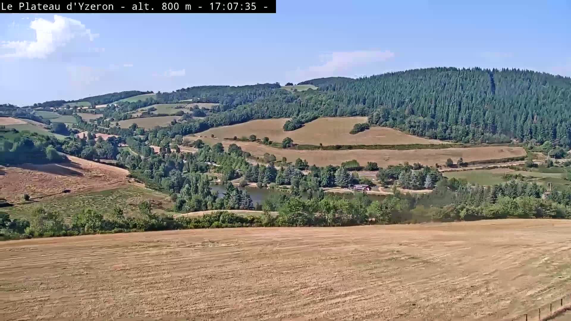 A sunny view from a hilltop shows a small village nestled in a valley surrounded by fields and forests, with a small structure and fenced area in the foreground.