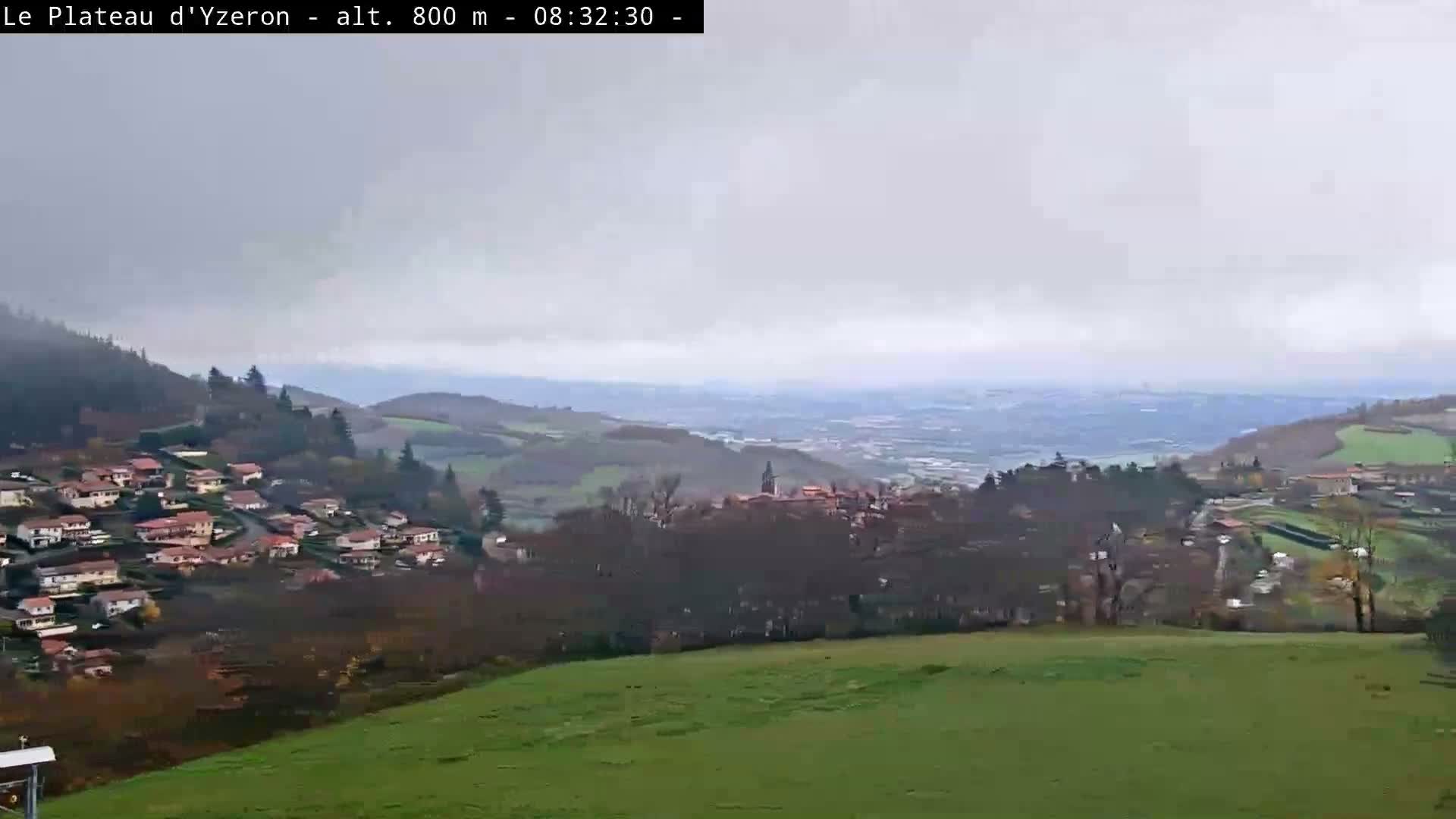 A cloudy day reveals a panoramic vista of rolling hills dotted with scattered villages and patches of green fields, all under a grey, overcast sky.