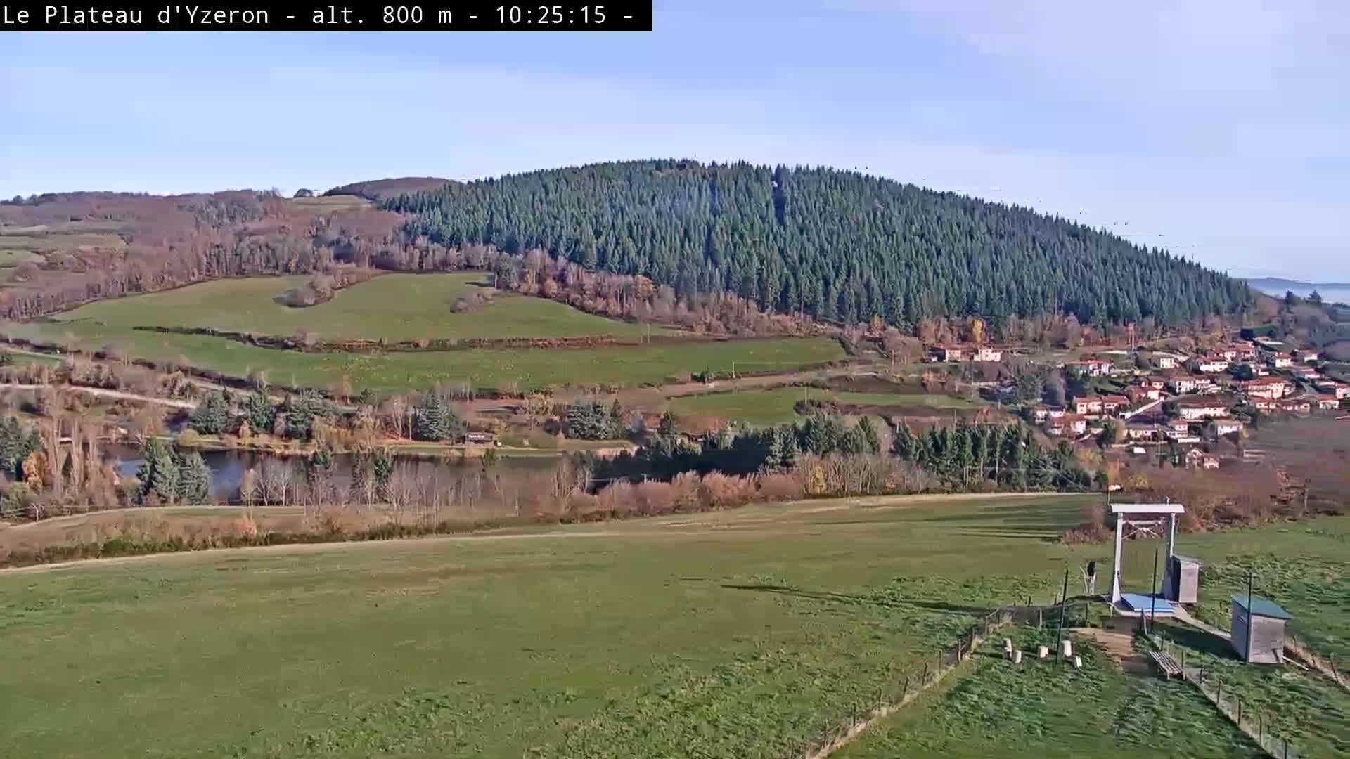 On a clear and sunny day, a rural landscape unfolds with a wide green field in the foreground where some geese are near a large wooden frame and small shelters, leading to a river, a village nestled against a densely forested hill, and more rolling terrain in the background.