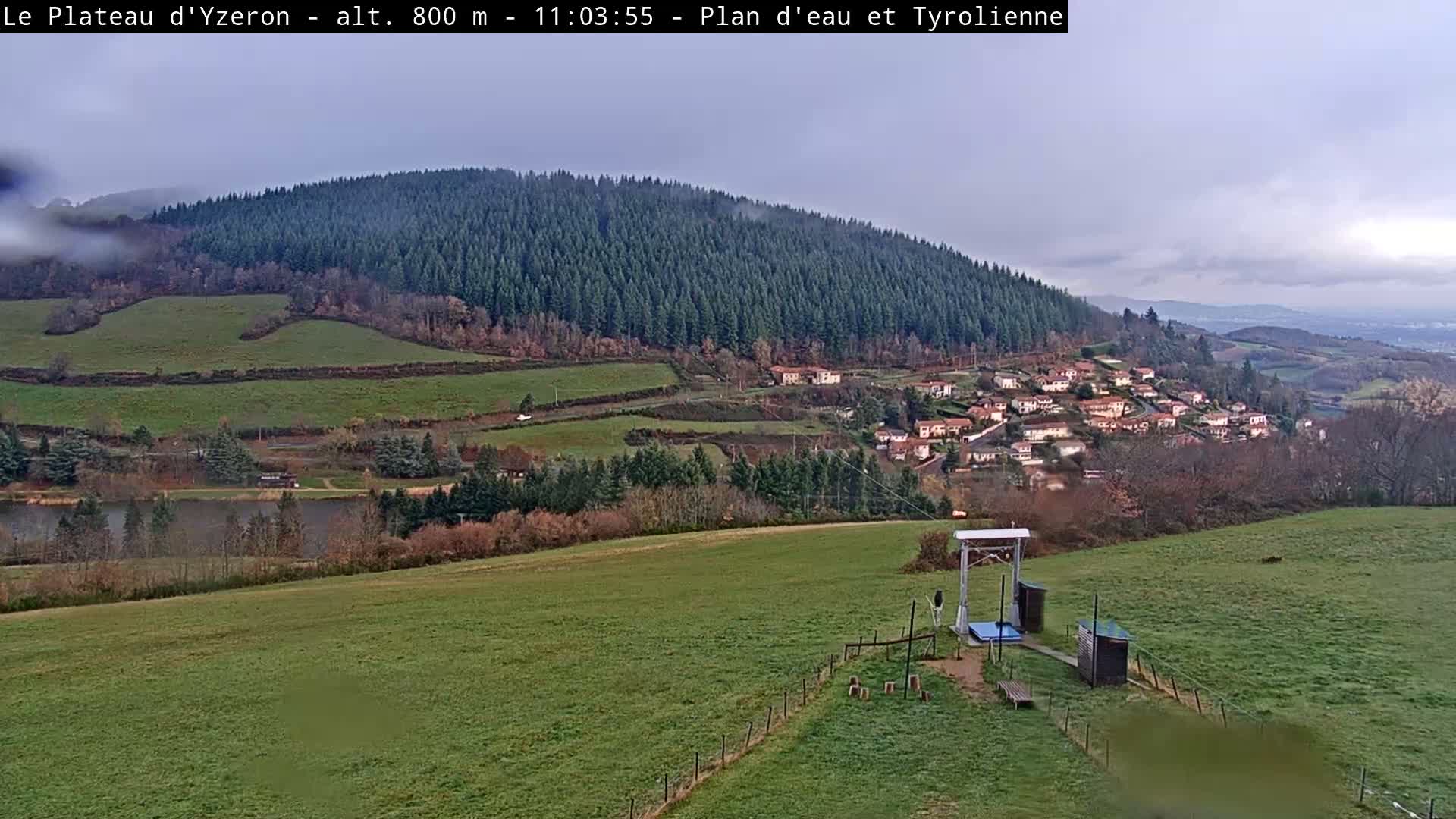 An expansive outdoor scene reveals a green, hilly landscape under an overcast and possibly misty sky, featuring a dark lake, a village clinging to a densely forested slope, and a visible zipline station in the foreground.