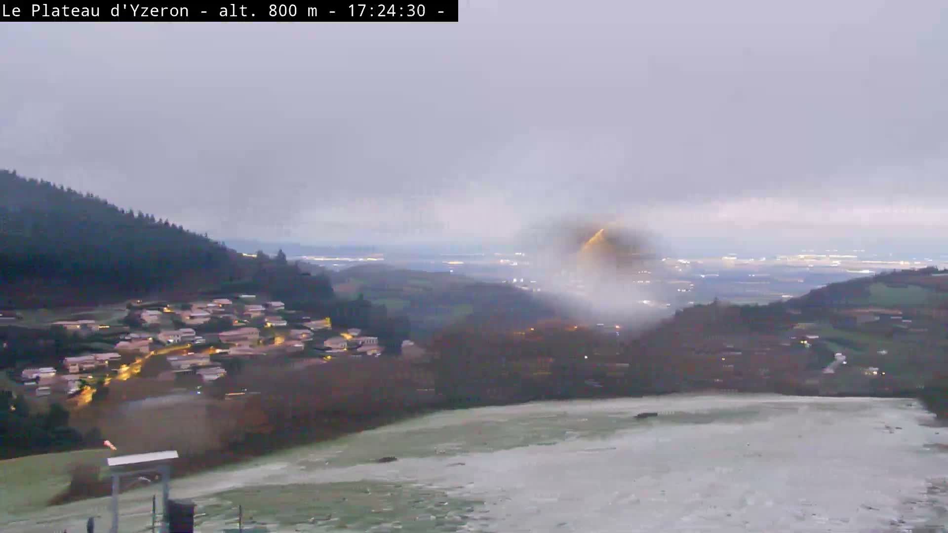 An expansive outdoor scene reveals a green, hilly landscape under an overcast and possibly misty sky, featuring a dark lake, a village clinging to a densely forested slope, and a visible zipline station in the foreground.
