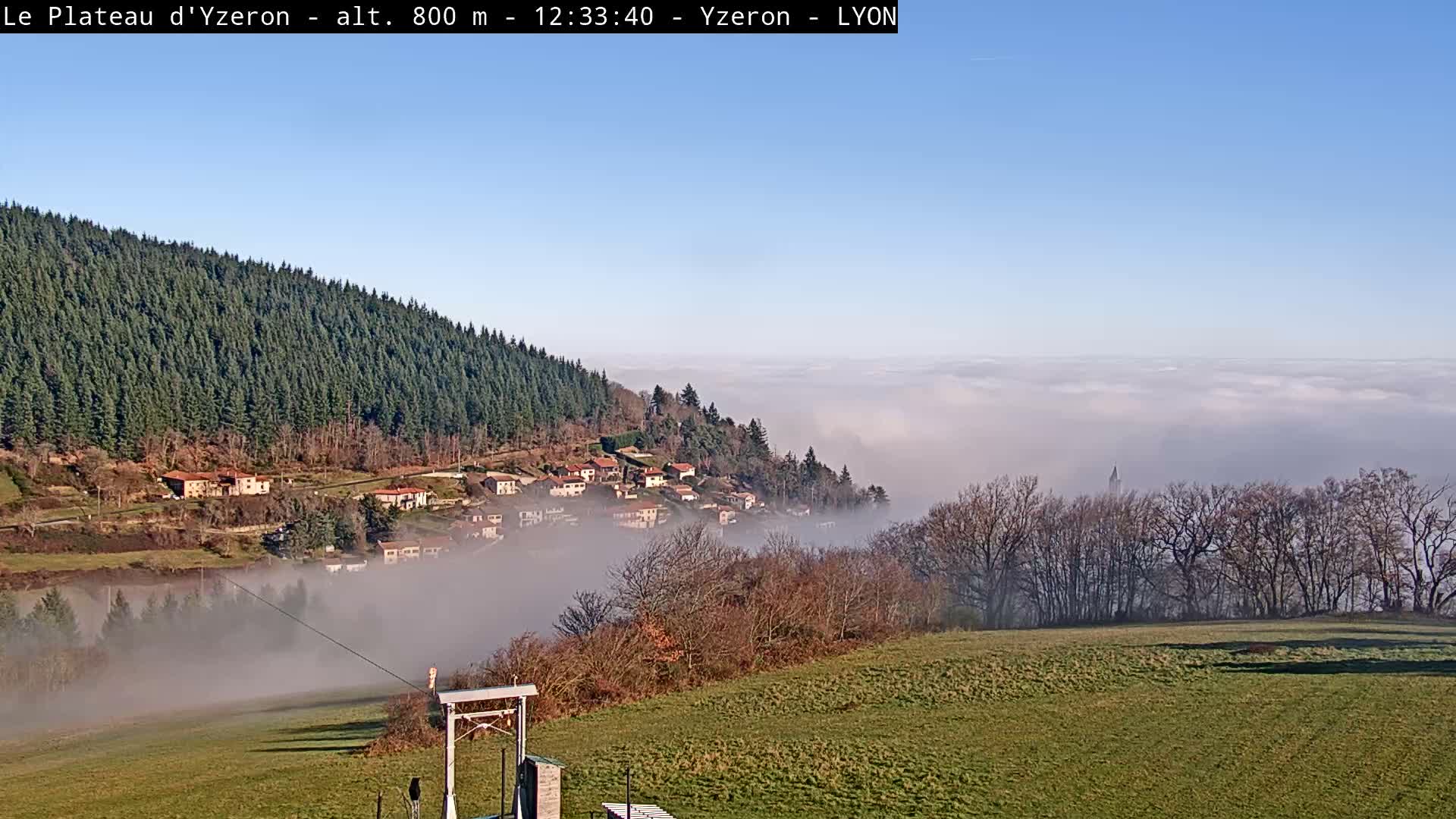 An expansive outdoor scene reveals a green, hilly landscape under an overcast and possibly misty sky, featuring a dark lake, a village clinging to a densely forested slope, and a visible zipline station in the foreground.