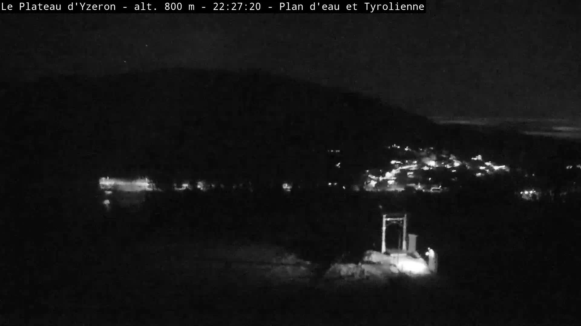 An expansive outdoor scene reveals a green, hilly landscape under an overcast and possibly misty sky, featuring a dark lake, a village clinging to a densely forested slope, and a visible zipline station in the foreground.