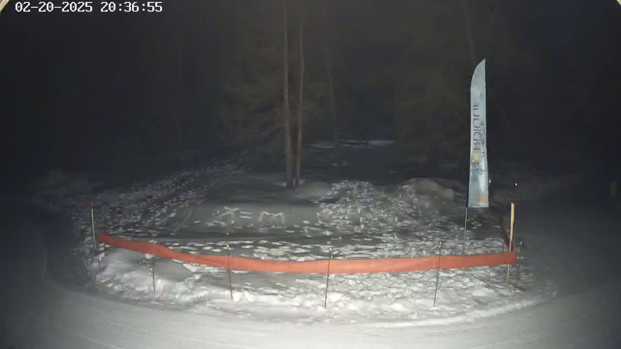 A snow-covered area at night is partially enclosed by an orange barrier, with a tall, slender sign visible in the background.