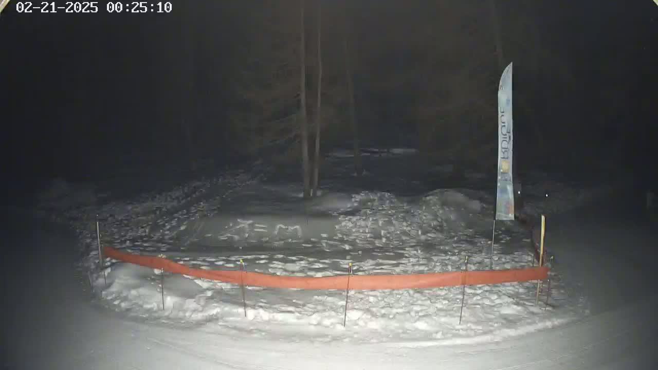 A snow-covered area at night, partially enclosed by an orange barrier, with a tall, thin banner standing near the center.