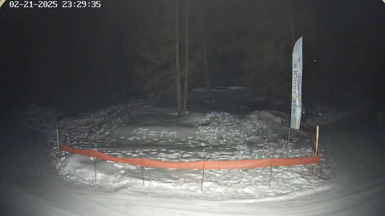 A snow-covered area at night, partially enclosed by an orange barrier, with a tall, thin banner visible in the background under dark, nighttime conditions.