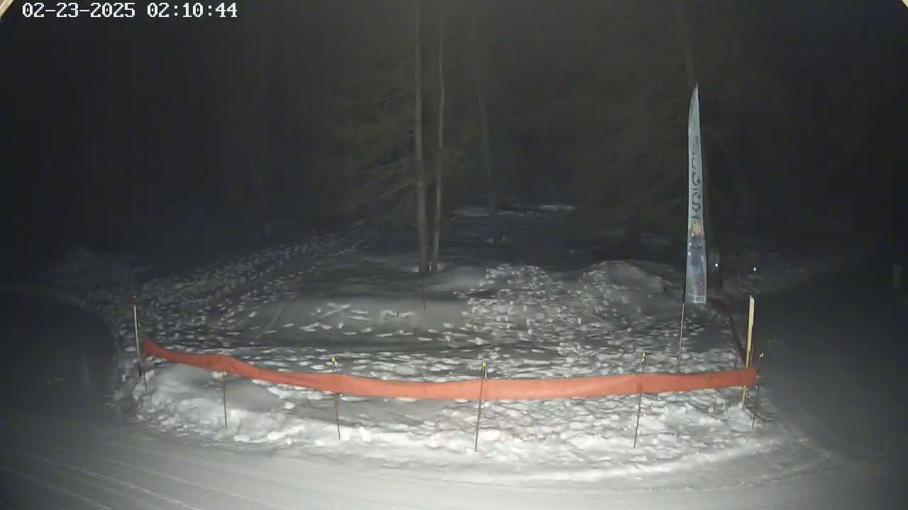 A snow-covered area at night, with a long orange barrier and a tall, thin signpost, is dimly lit under dark skies.