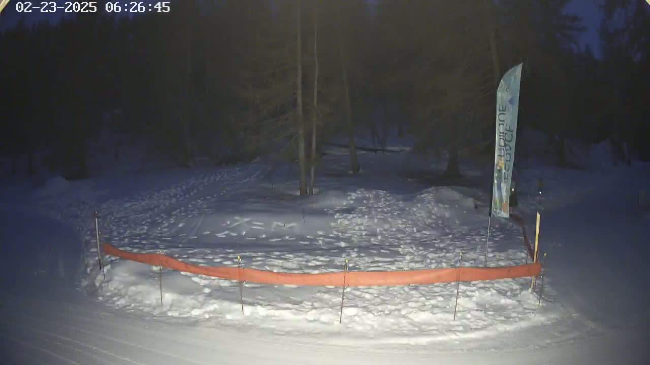 A snow-covered area at night, bordered by a low orange fence, is partially surrounded by dark trees.