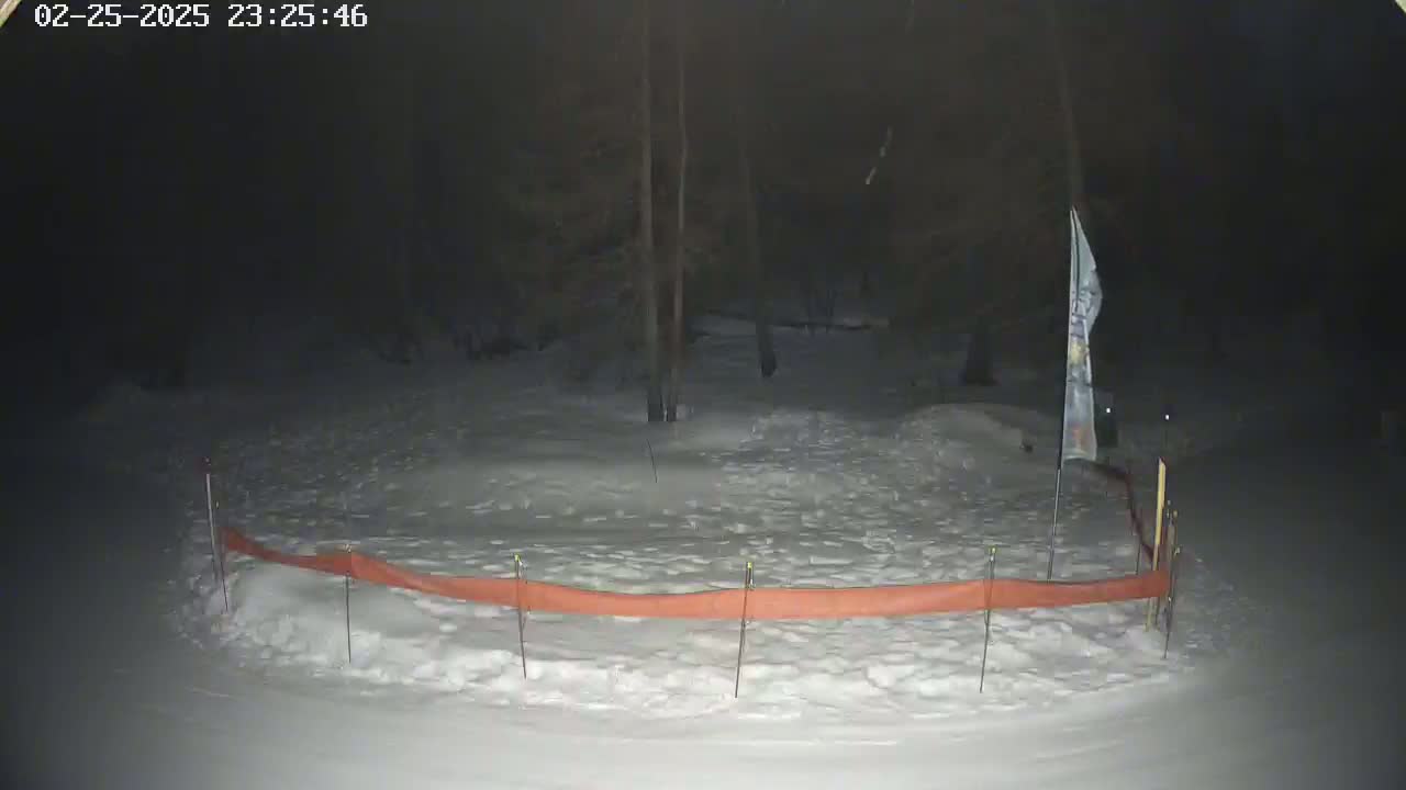 A snow-covered area at night, partially enclosed by an orange barrier, with trees and a flag visible in the background.