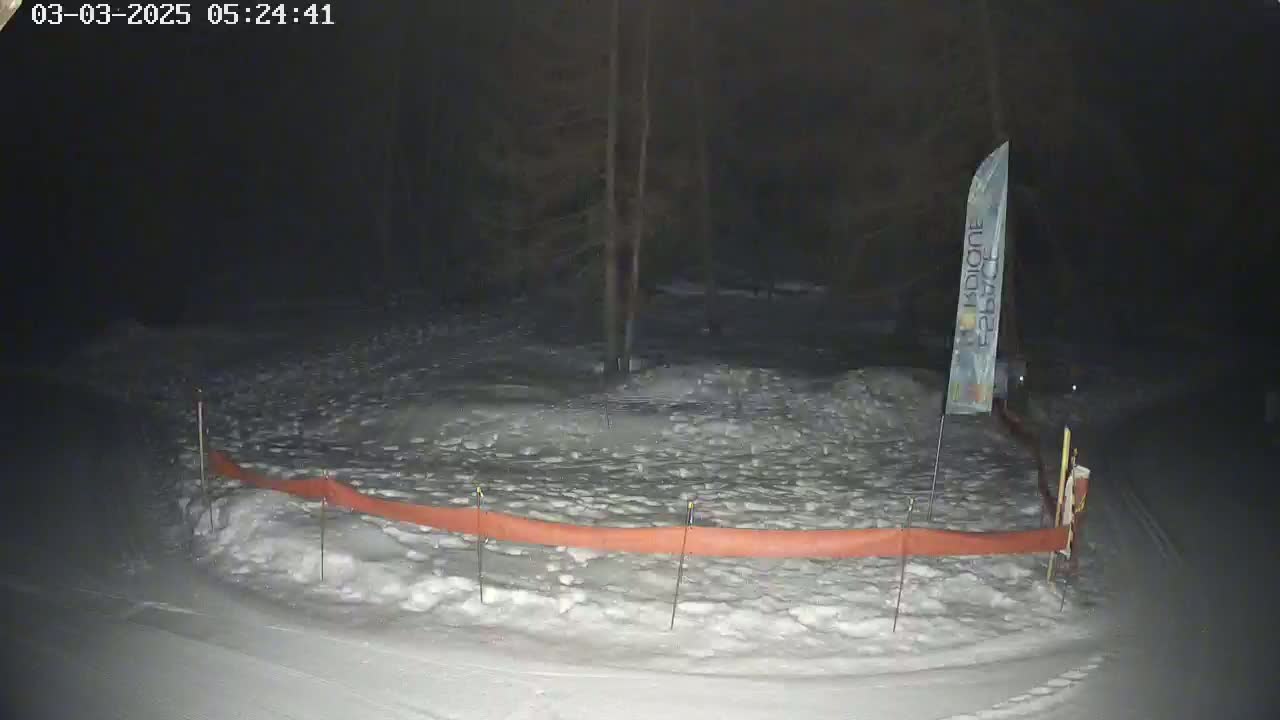A snow-covered area at night is enclosed by an orange barrier, with a tall, thin banner standing nearby.
