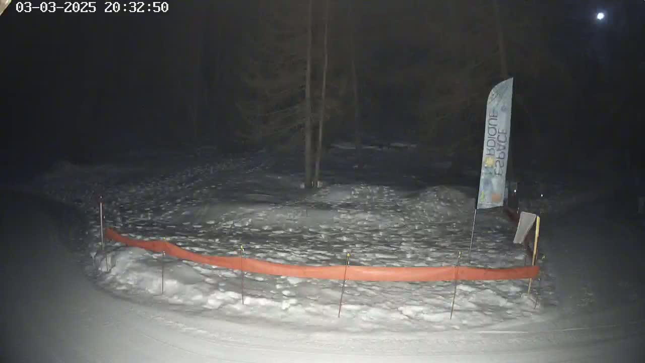 A snow-covered area at night, illuminated by a moon, features a low orange barrier and a single flagpole.