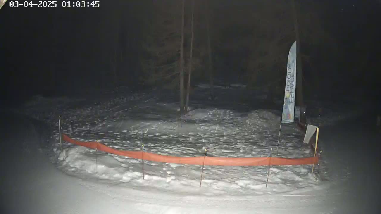 A snow-covered area at night, illuminated by a faint light source, features a long orange barrier and a tall vertical banner.