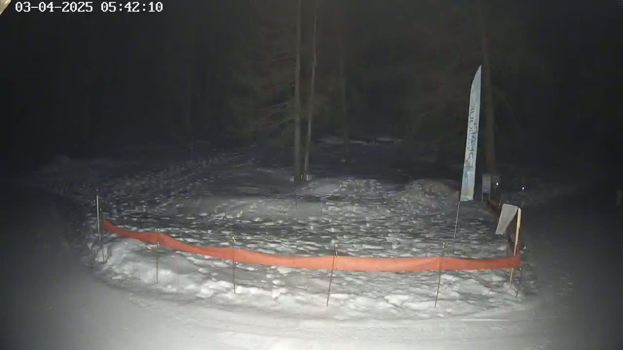 A snow-covered area at night, partially enclosed by an orange barrier, is dimly lit near a dark wooded area.
