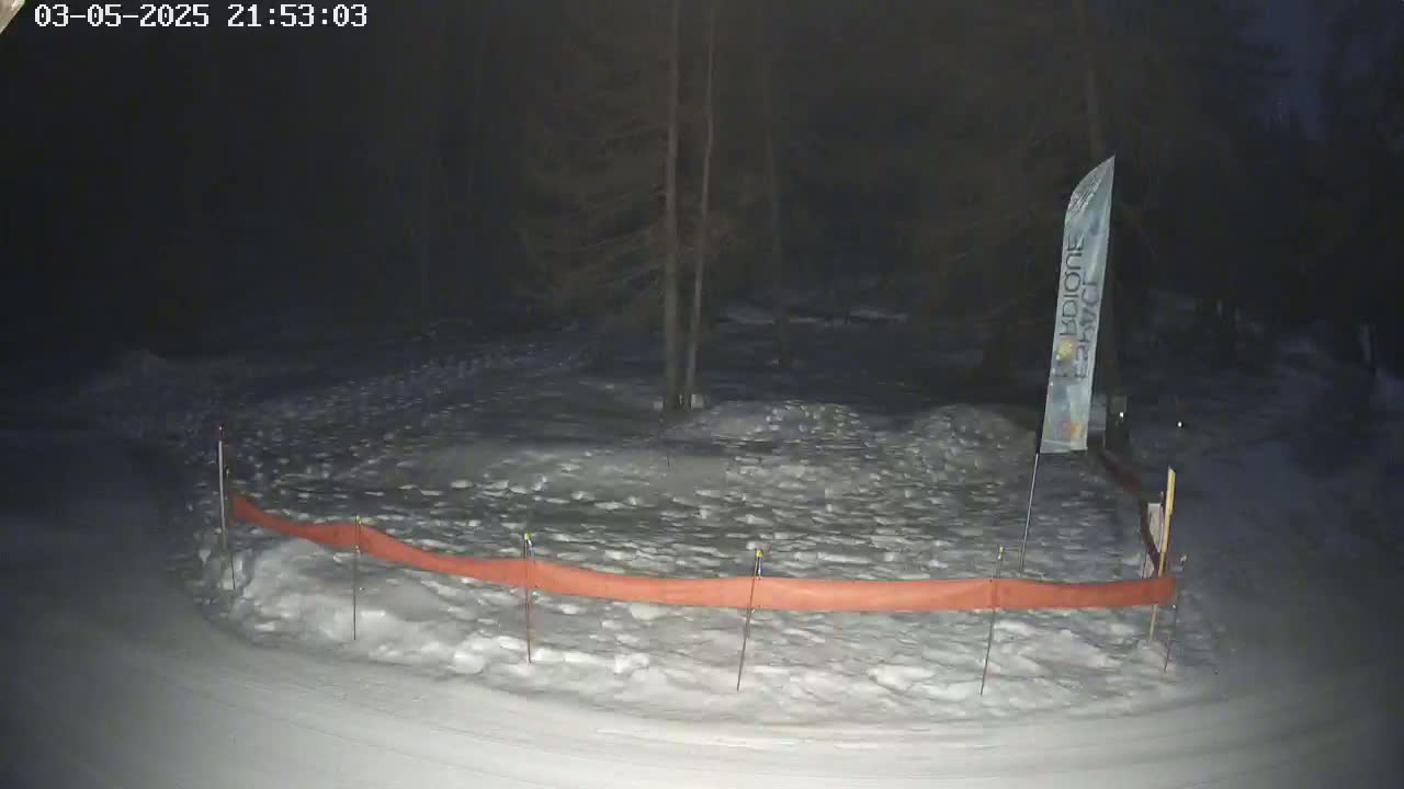 A snow-covered area at night, enclosed by an orange barrier, with trees in the background under dark conditions.