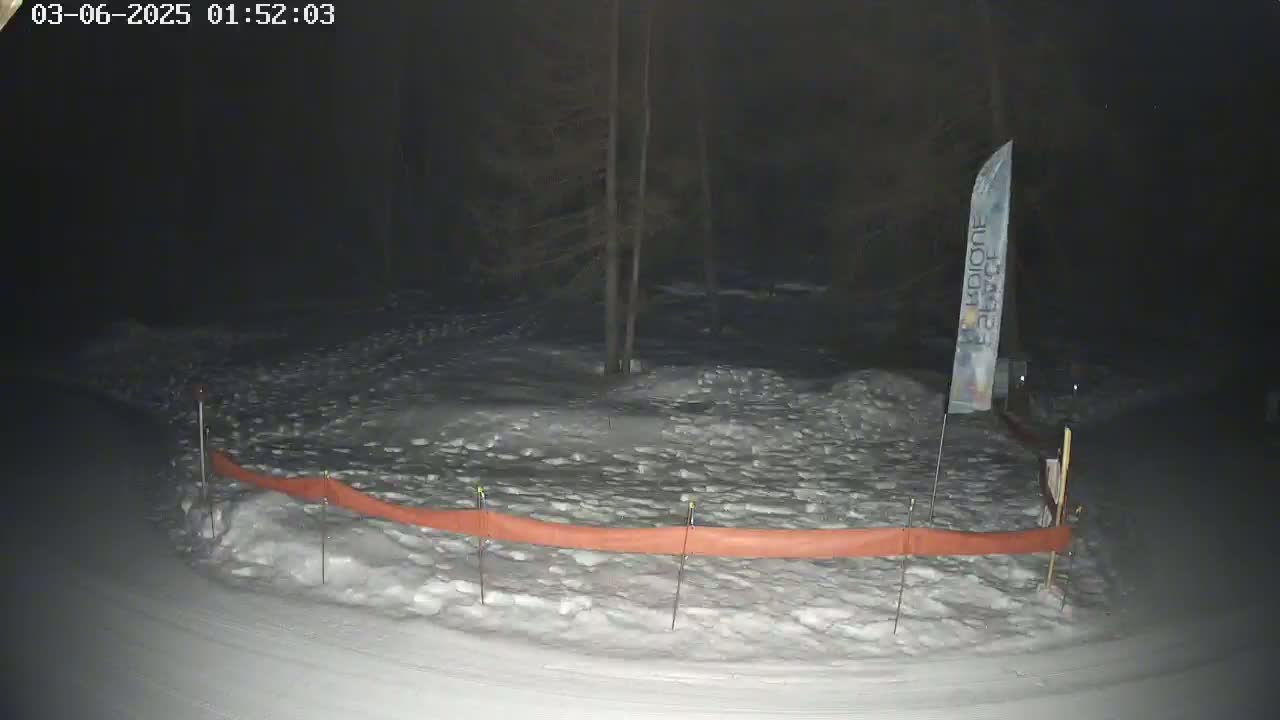 A snow-covered area at night, bordered by an orange snow fence and a tall banner, is illuminated by a faint light source.